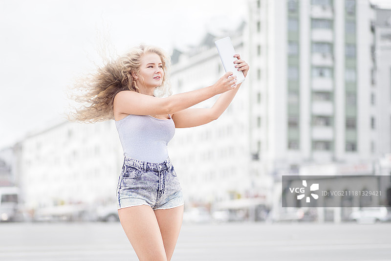 年轻女子脖子上戴着复古音乐耳机，用平板电脑自拍，在城市背景下微笑图片素材