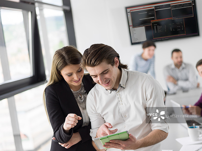 两位商务人士使用平板电脑为下次会议做准备，并在背景中与同事讨论想法图片素材
