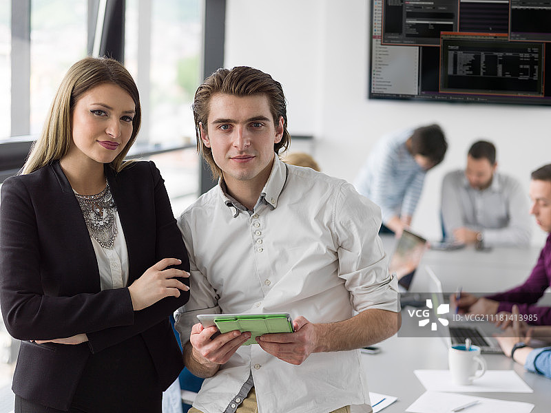 两位商务人士使用平板电脑为下次会议做准备，并在背景中与同事讨论想法图片素材