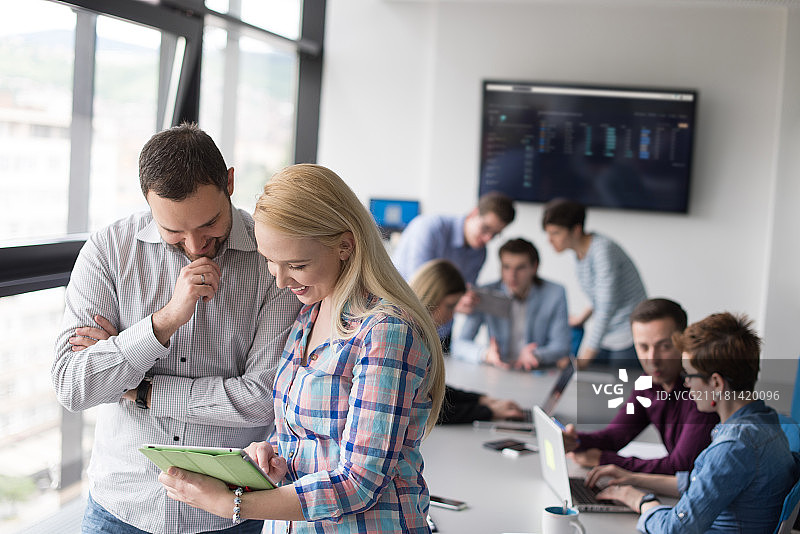 两位商务人士使用平板电脑为下次会议做准备，并在背景中与同事讨论想法图片素材