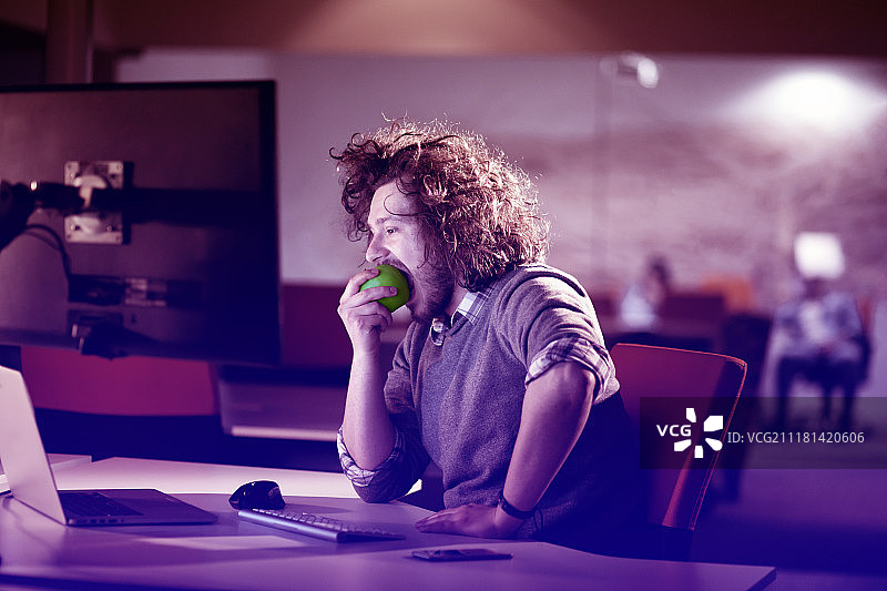 富有创意的男人在夜晚的办公室里工作并吃苹果图片素材
