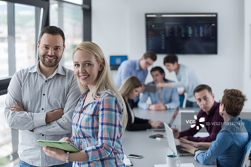 两位商务人士使用平板电脑为下次会议做准备，并在背景中与同事讨论想法图片素材