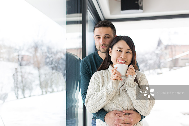快乐的年轻多元民族情侣在寒冷的冬日在家中靠窗享用早咖啡图片素材