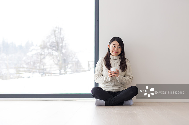在寒冷的冬日早晨，年轻漂亮的亚洲女性在窗边地板上享受咖啡图片素材