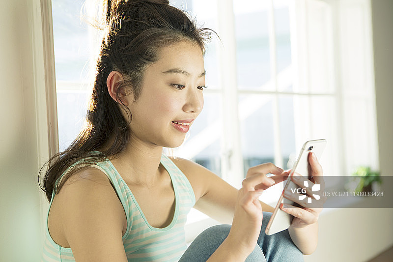 微笑的年轻女人在窗边使用智能手机图片素材