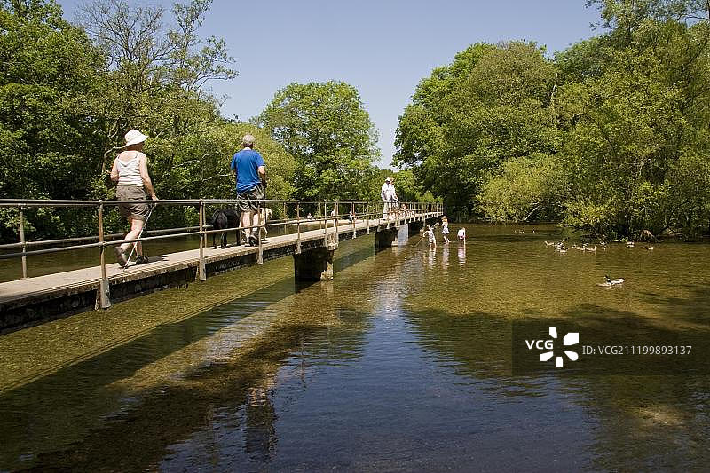 人们走过狭窄的人行桥,孩子们在浅河里玩耍,弗罗姆河,莫雷顿,多塞特,英格兰,英国,欧洲图片素材