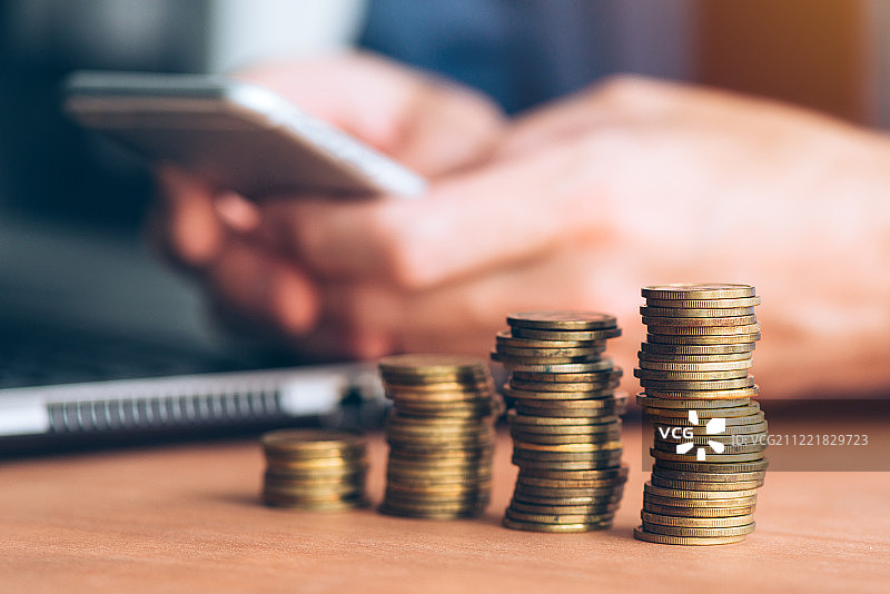 前景中堆叠着硬币，使用智能手机的商人图片素材