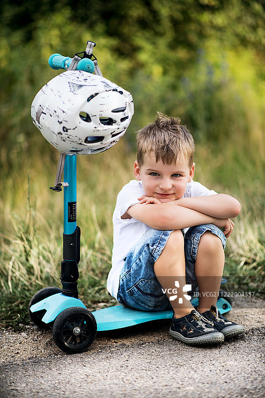 一个戴着头盔的小男孩在夏日公园的道路上坐在滑板车上图片素材