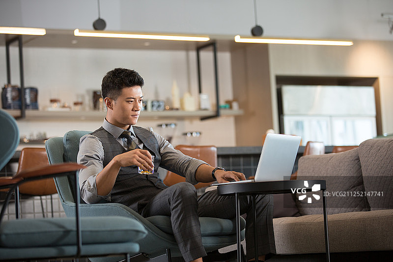 商务青年男子在酒店休闲区喝酒使用笔记本电脑工作图片素材