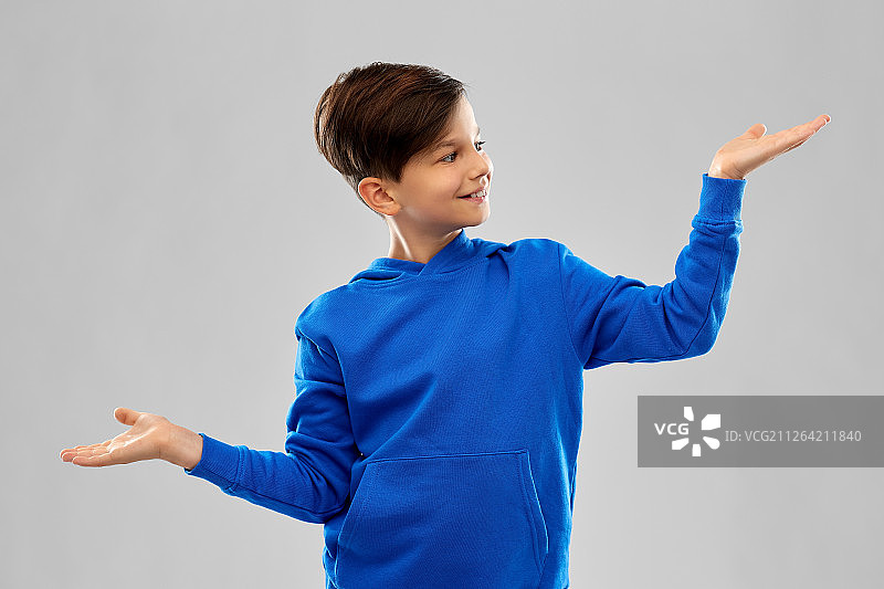 儿童、手势和人物概念 - 灰色背景上空手拿着想象东西的蓝色连帽衫微笑男孩图片素材