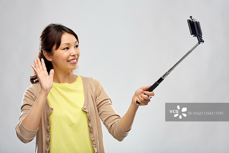 科技与人：快乐的亚洲女人用自拍杆和智能手机拍照，在灰色背景上挥手。图片素材