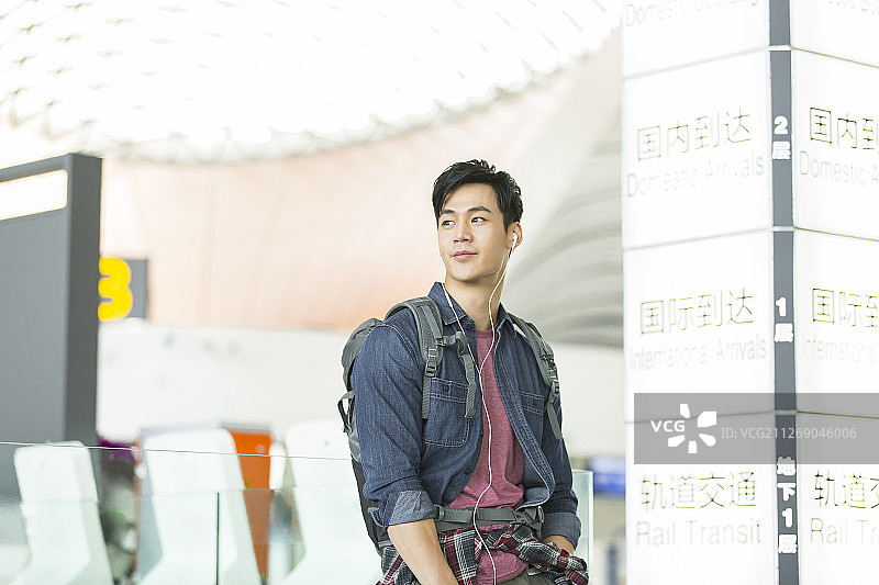 青年男子背着旅行包使用手机戴耳机听音乐在机场休息放松等候飞机起飞图片素材