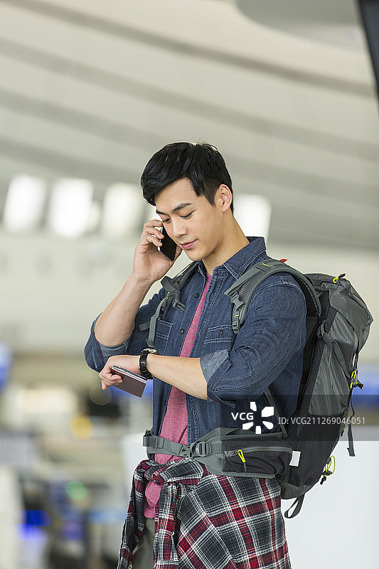 青年男子背着旅行包使用手机打电话拿着护照和机票看手表核实时间在机场图片素材