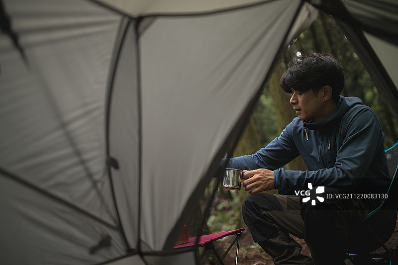 男人在森林里坐在帐篷旁边的椅子上喝咖啡的露营摄影照片图片素材