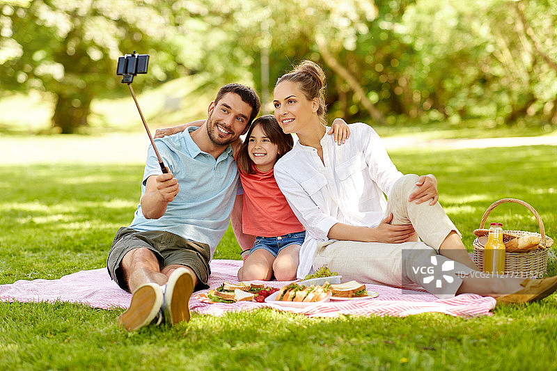 在夏季公园里，快乐的家庭野餐并用自拍杆通过智能手机拍照图片素材