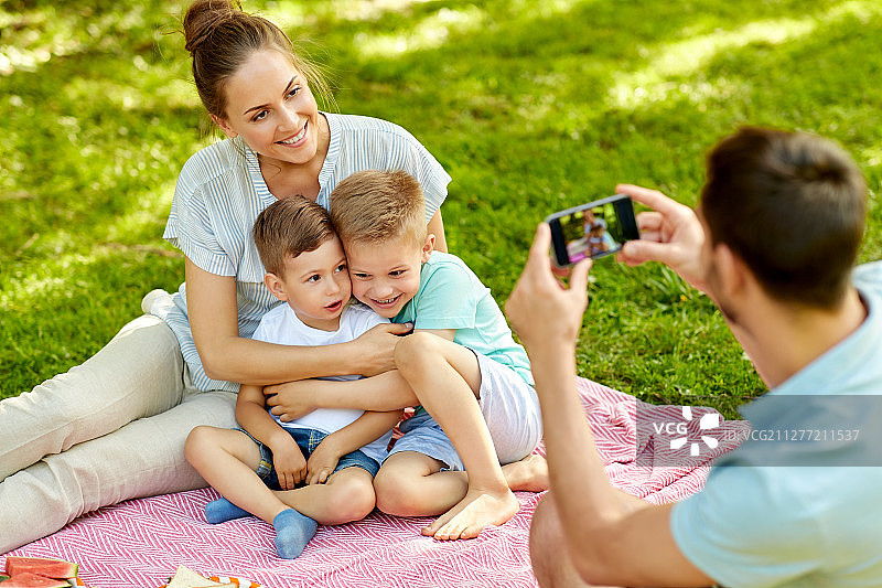 夏日公园野餐，父亲用智能手机给快乐的母亲和两个小儿子拍照图片素材
