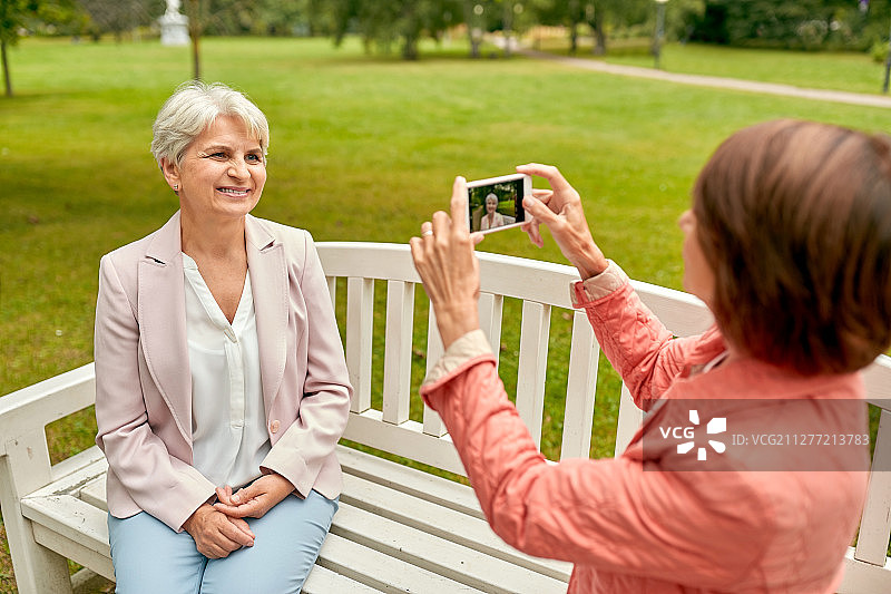 老年，退休和人们的概念-妇女拍摄她的朋友坐在夏季公园长椅上的智能手机。一位女士在公园里给她的朋友拍照图片素材