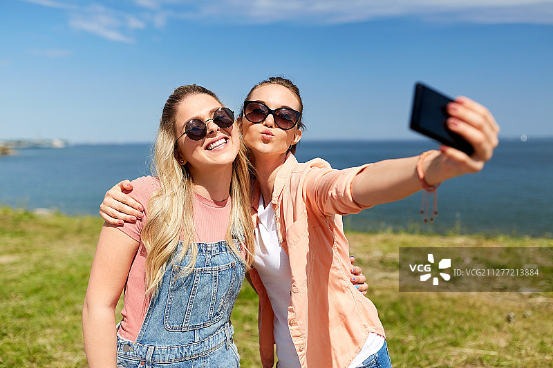 休闲友谊概念-快乐微笑的少女或最好的朋友戴着太阳镜在海边拥抱并用智能手机自拍图片素材