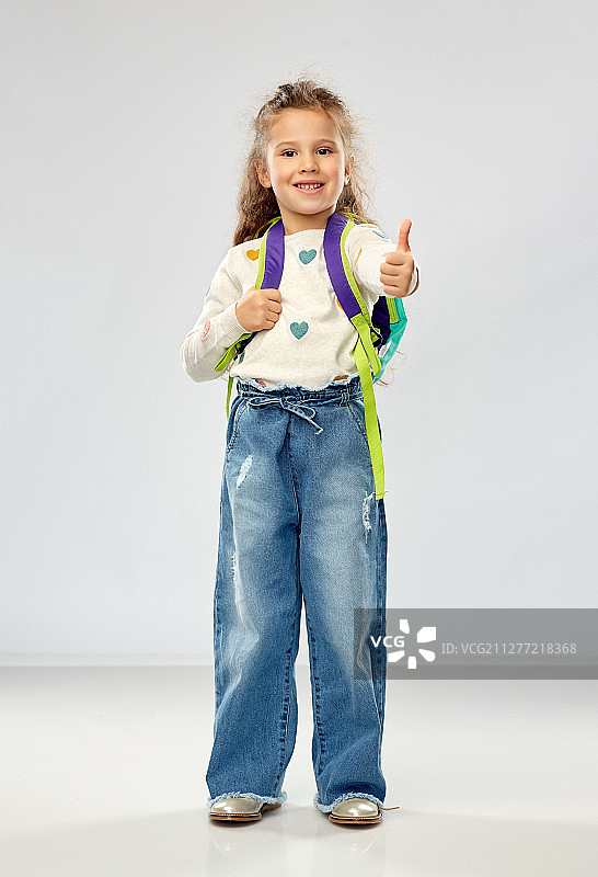 童年、学校和教育的概念-快乐的小女孩带着背包在灰色背景上竖起大拇指。快乐的女孩带着书包竖起大拇指图片素材