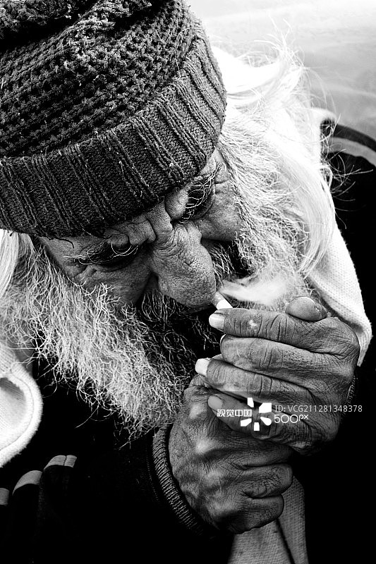 戴着无檐小便帽的单色胡须流浪汉点燃香烟图片素材