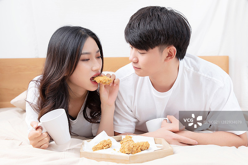 穿着白色短袖趴在床上的一对亚洲年轻情侣，正在快乐的吃着炸图片素材