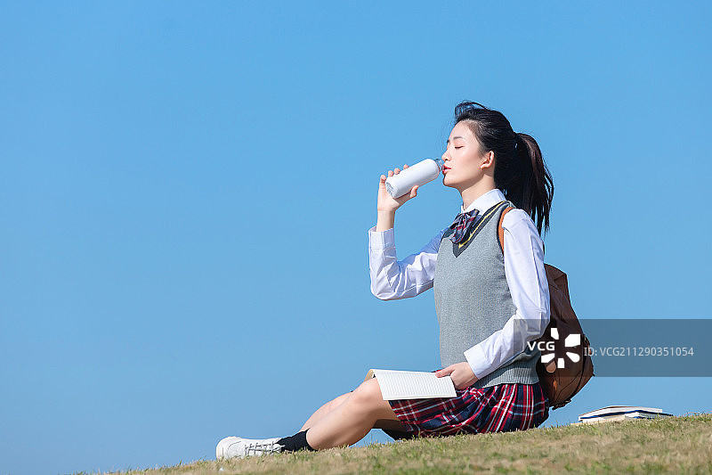 蓝色天空背景下，坐在草地上的亚洲女学生图片素材