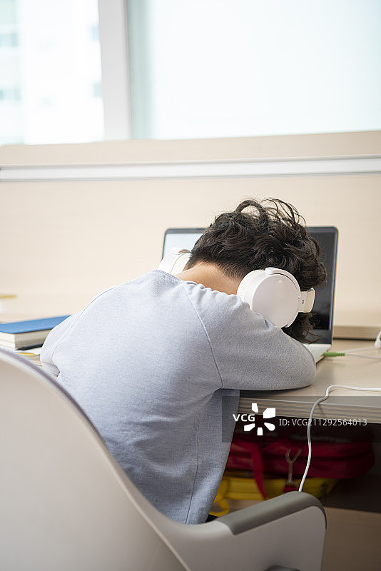 戴着耳机的男孩坐在书桌旁，趴着睡觉的摄影图片素材