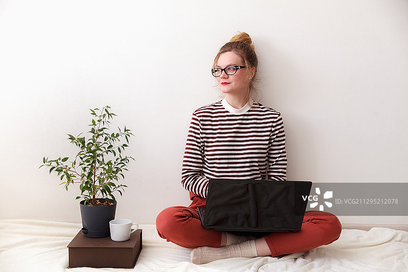 坐在家中（俄罗斯莫斯科）的床上使用笔记本电脑的年轻女人，有盆栽植物图片素材