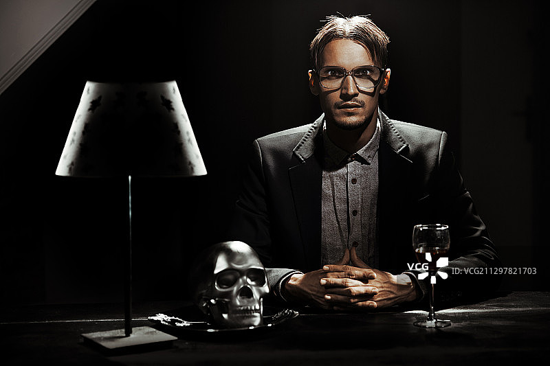 戴眼镜的反常男子或反社会者坐在桌旁，桌上放着一个人类头骨，适合做书籍封面图片素材