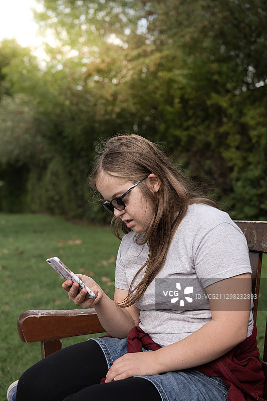 患有唐氏综合征的少女坐在公园的板凳上使用手机图片素材