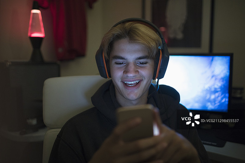微笑的青少年男孩戴着耳机在黑暗的房间里使用智能手机图片素材