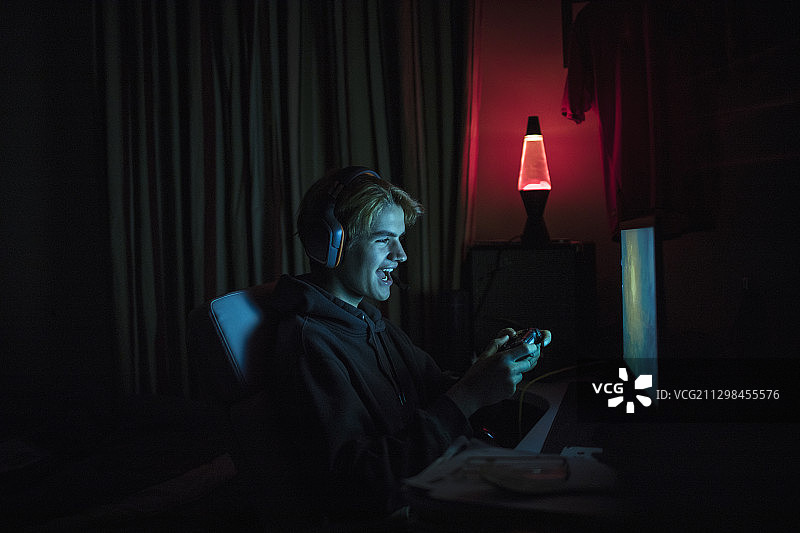 微笑的少年在黑暗卧室里用电脑玩电子游戏图片素材