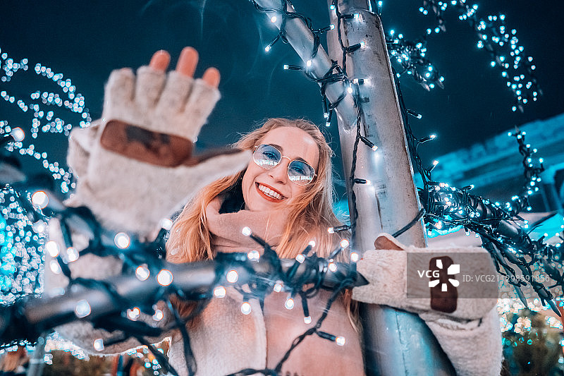 戴着太阳镜站在灯光闪烁的圣诞树旁的微笑女人图片素材