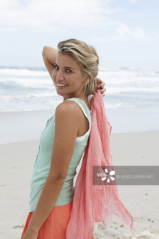 一位金发女人在海滩上，戴着围巾，穿着绿松石色上衣和鲑鱼粉色裙子图片素材