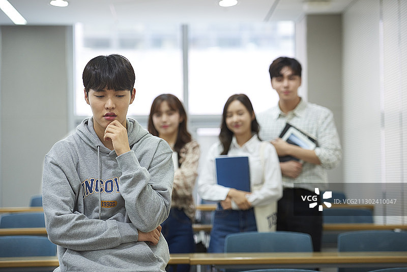 大学生坐在书桌旁，表情担忧，三名学生站在讲堂后方图片素材