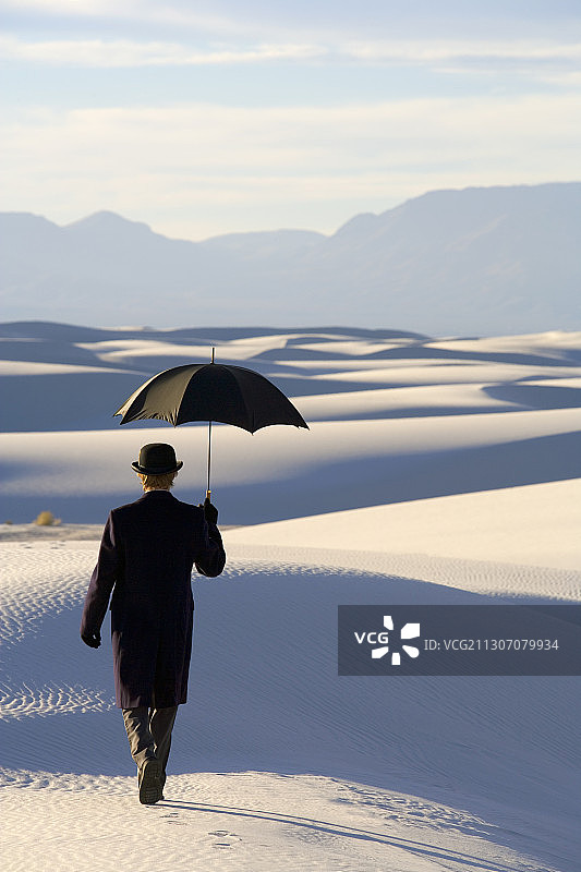 身穿黑外套和西装，头戴圆顶礼帽，手持雨伞的男人站在白色沙漠中图片素材
