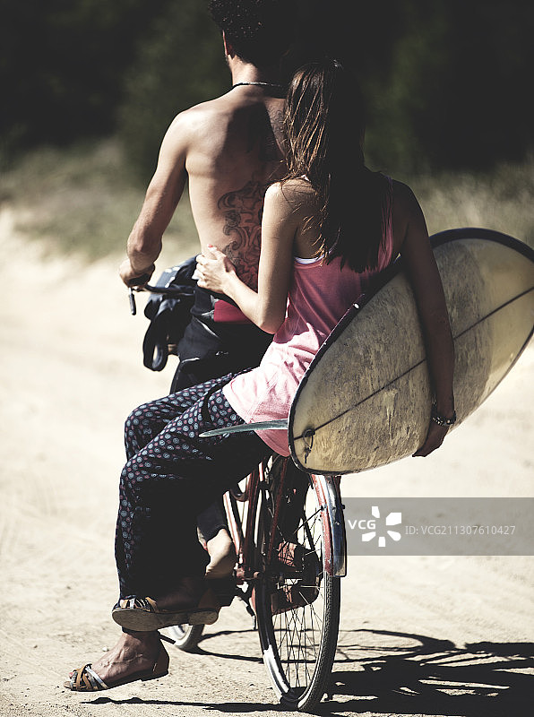男子骑自行车，女子坐在后座上抱着冲浪板的背影图片素材
