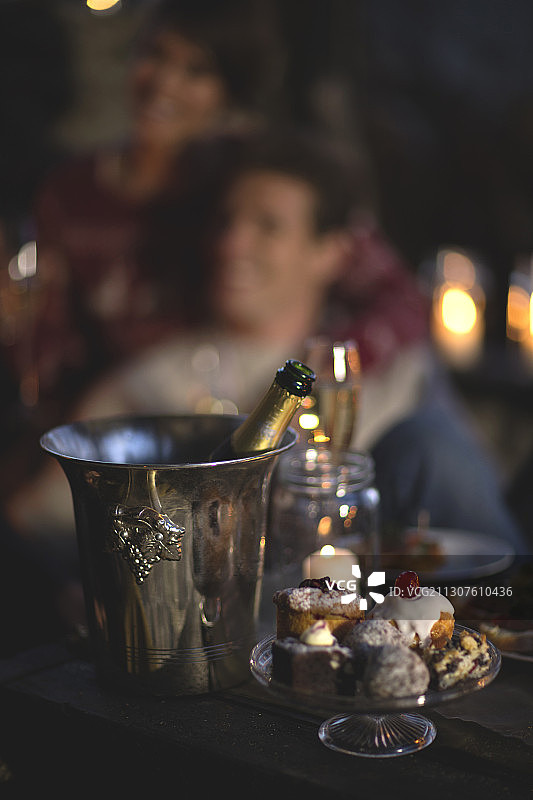 香槟酒瓶置于金属冰桶中，玻璃蛋糕架上摆放着各式蛋糕，背景中有人图片素材