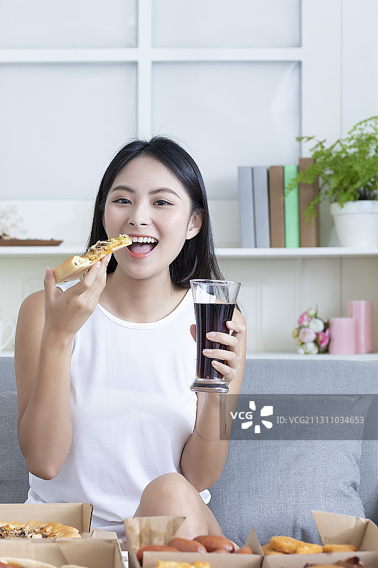 东方美女坐在沙发上一手拿着可乐一手拿着披萨正在吃图片素材