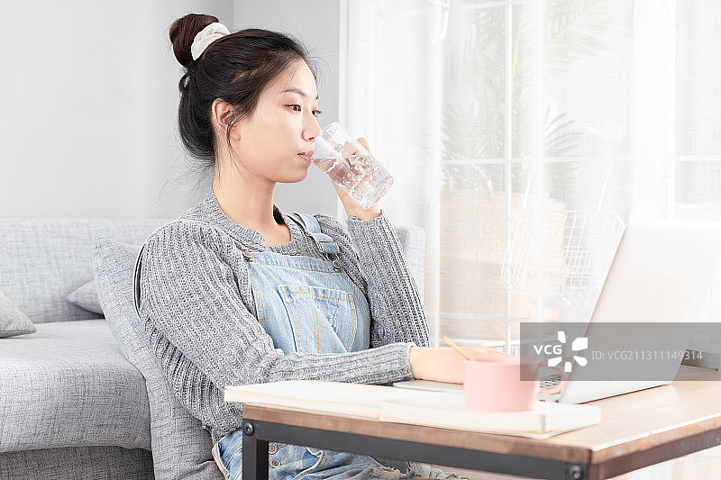 坐在地毯上，正在上网课并喝水的亚洲女孩图片素材