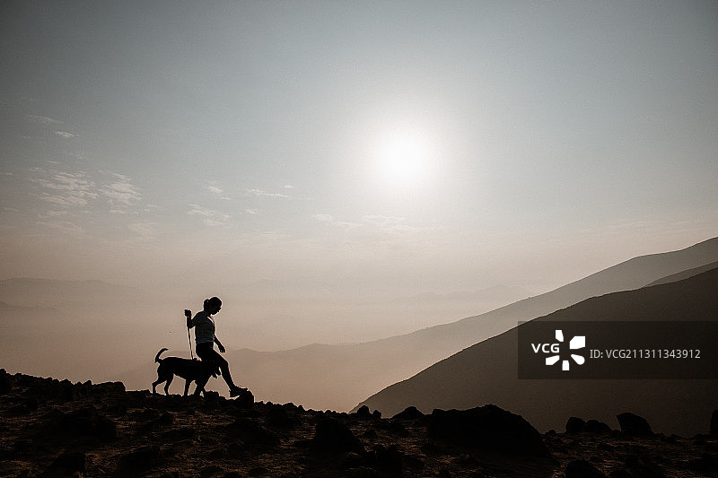 日落时分站在岩石上与狗对视的剪影女人，秘鲁图片素材