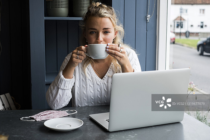 沃灵顿，英格兰，年轻的金发女人独自坐在咖啡馆的桌子旁，带着笔记本电脑和一杯咖啡图片素材