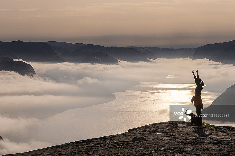 女人和狗站在布道石上，比出和平手势，身后是挪威吕瑟峡湾的日出美景图片素材