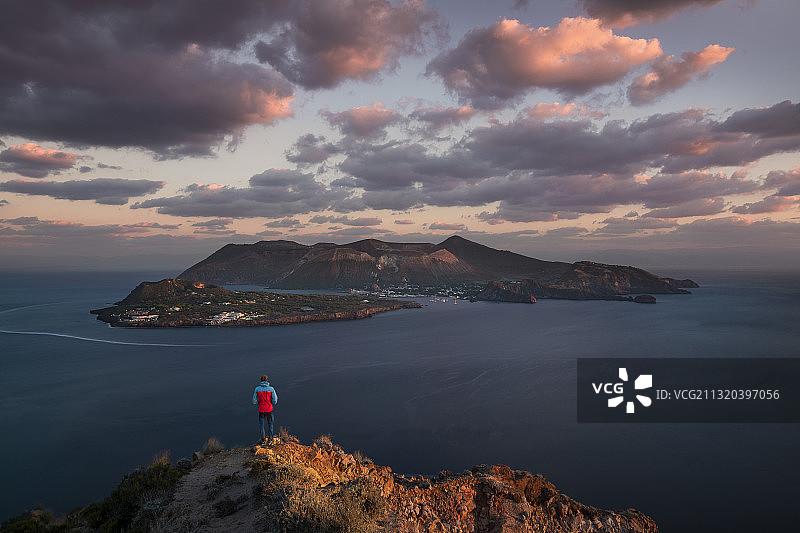 身穿红色夹克的男人站在利帕里海岸，眺望日落时分的火山岛，意大利西西里岛图片素材