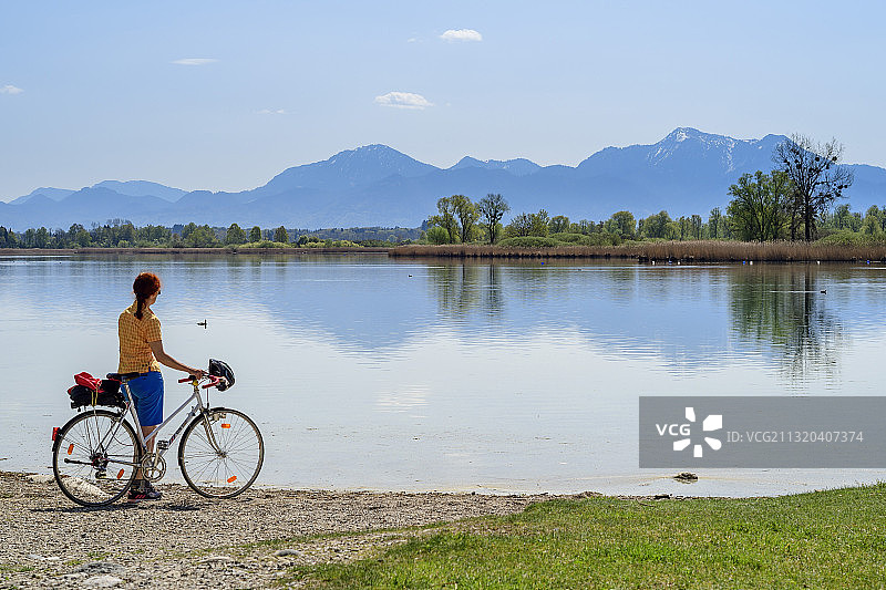 女人骑自行车站在岸边，眺望基姆湖、霍赫费尔恩山和霍赫格恩山，德国巴伐利亚州上巴伐利亚基姆高基姆湖自行车道图片素材