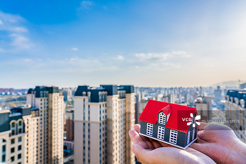 楼房背景下手托着一个小房子玩具——买房子概念图片素材