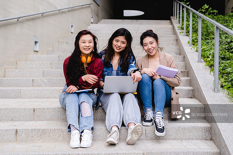 坐在楼梯上使用笔记本电脑的微笑朋友们肖像图片素材