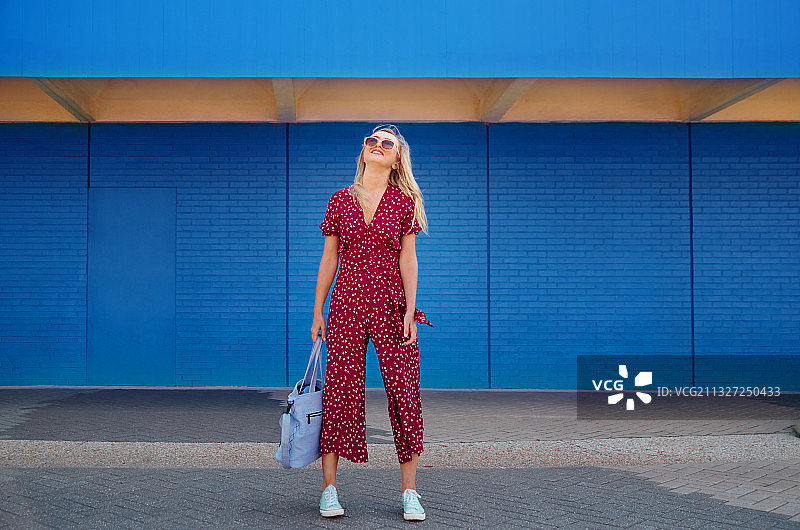 穿着勃艮第连身裤、戴着太阳镜的漂亮金发女人，靠在蓝色墙上，拿着天蓝色手提袋图片素材