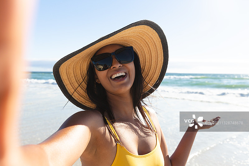 戴着遮阳帽和太阳镜在海滩上自拍的混血微笑女性图片素材
