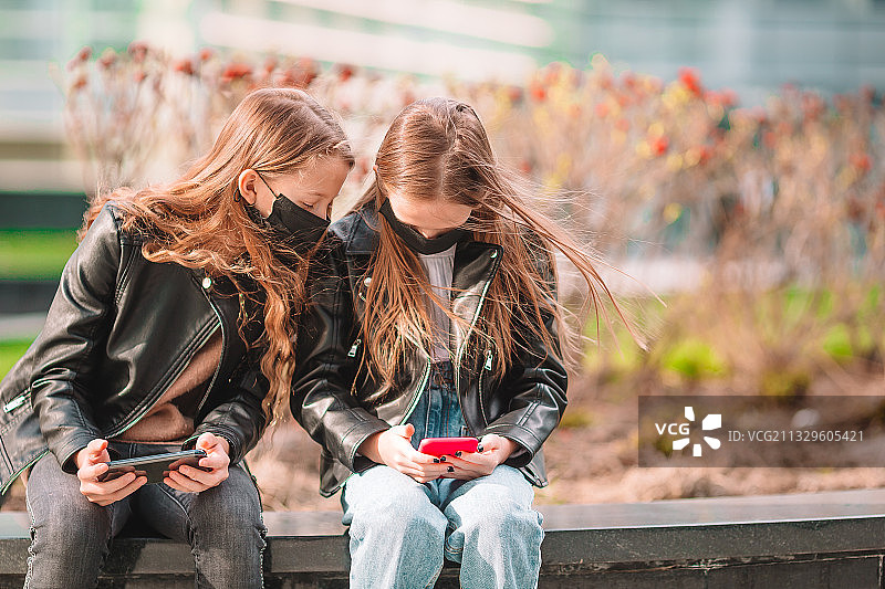 两个女孩戴着口罩坐在城市的长椅上使用手机图片素材